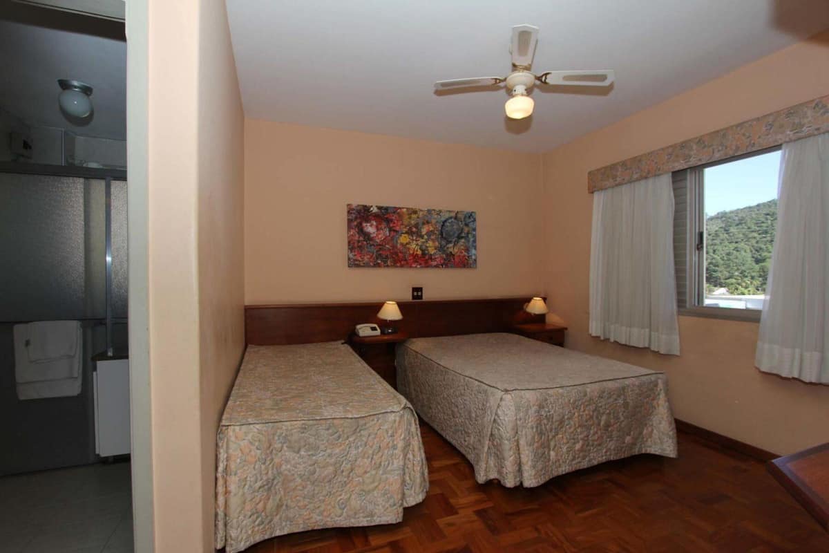 Ein Zimmer mit zwei Einzelbetten, einem Deckenventilator, einem Bild an der Wand und einem Fenster mit Vorhängen.