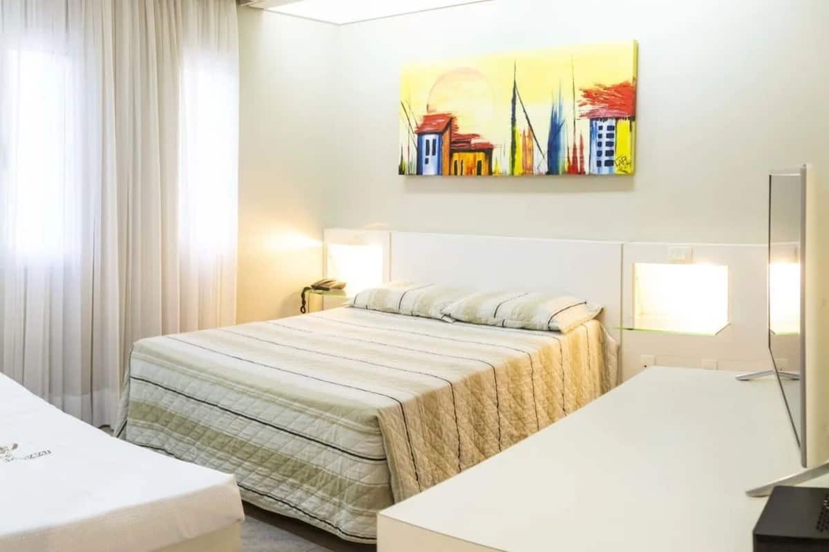 Ein Hotelzimmer mit Bett, Schreibtisch, Computer, einem Gemälde an der Wand und einem Badezimmer mit Dusche und Toilette.