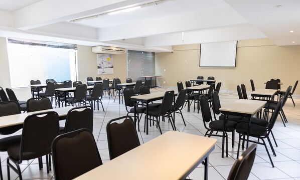 Un salón de conferencias con varias mesas y sillas, una pantalla de proyector y un ventanal con persianas.