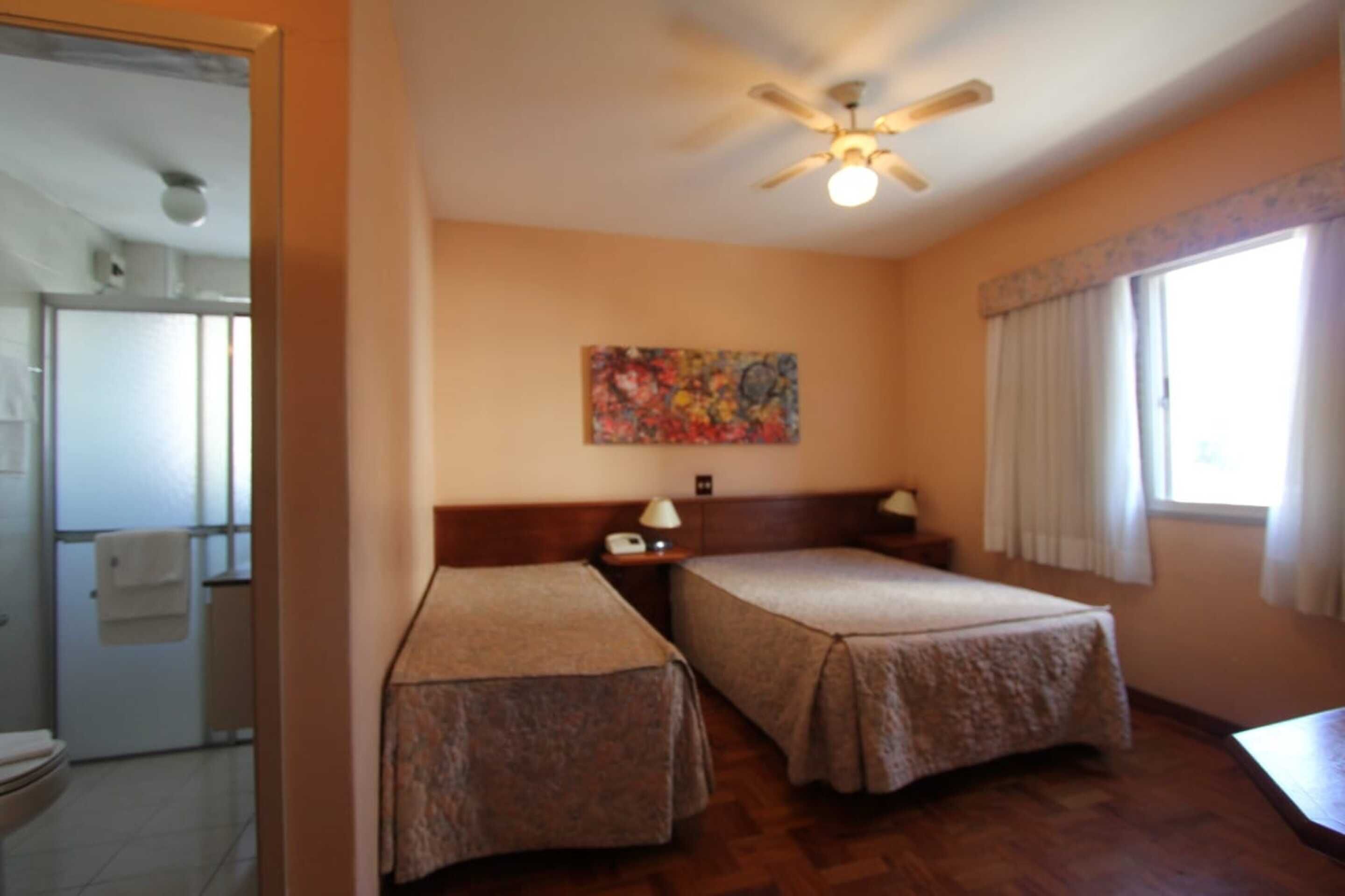 A hotel room with two beds, a ceiling fan, a painting on the wall, and a window with curtains.