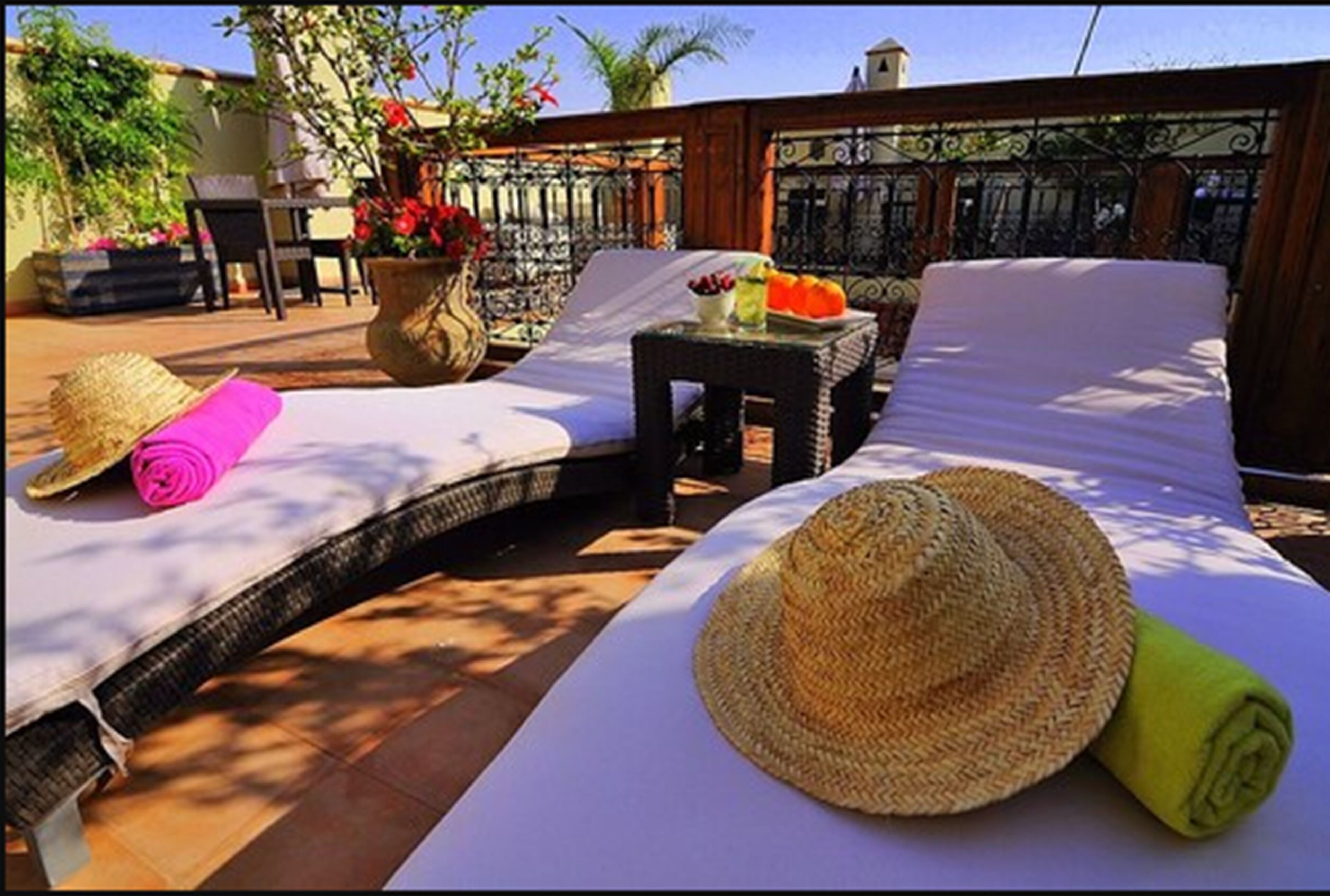 A sunlit outdoor lounge area with two lounge chairs, a small table with drinks, and a large woven hat on the ground.