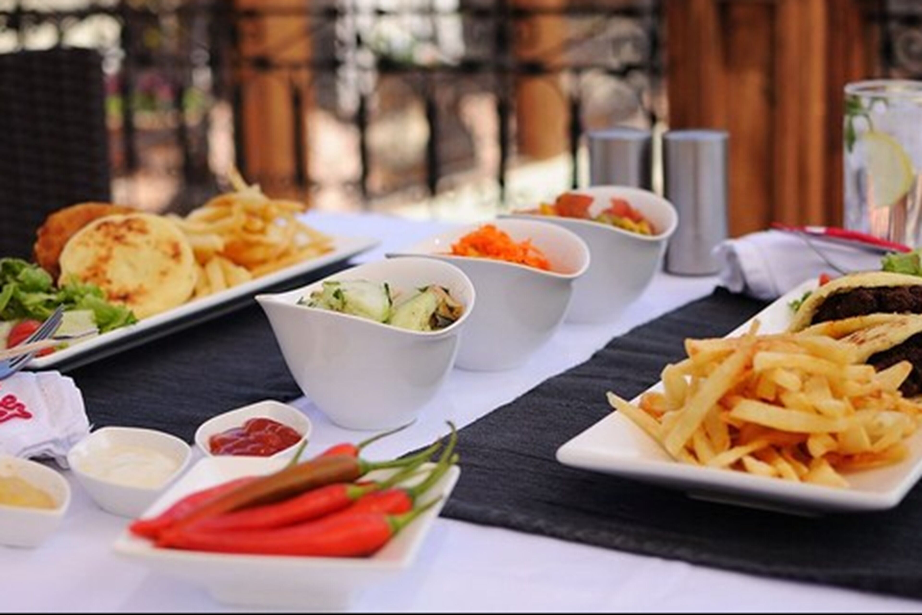 Ein Tisch ist reichhaltig mit Speisen gedeckt, darunter Pommes, Salate und ein Burger, mit Soßen und einem Getränk im Hintergrund.