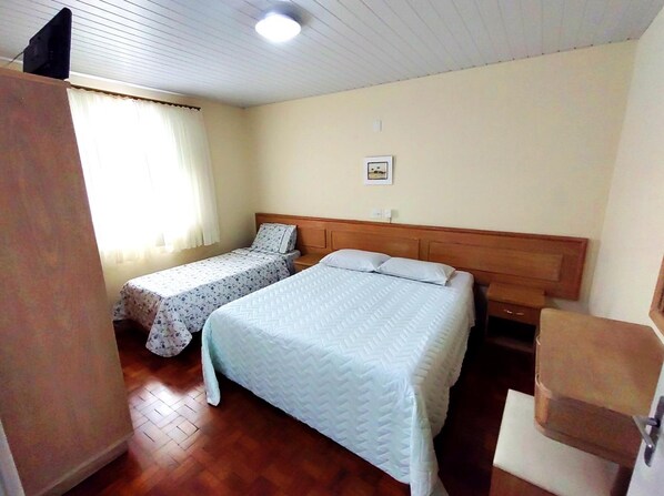 A room with two beds, a wooden headboard, a small bedside table, a chair, and a window with curtains. - Pousada Águas de Lindóia (Águas de Lindóia)