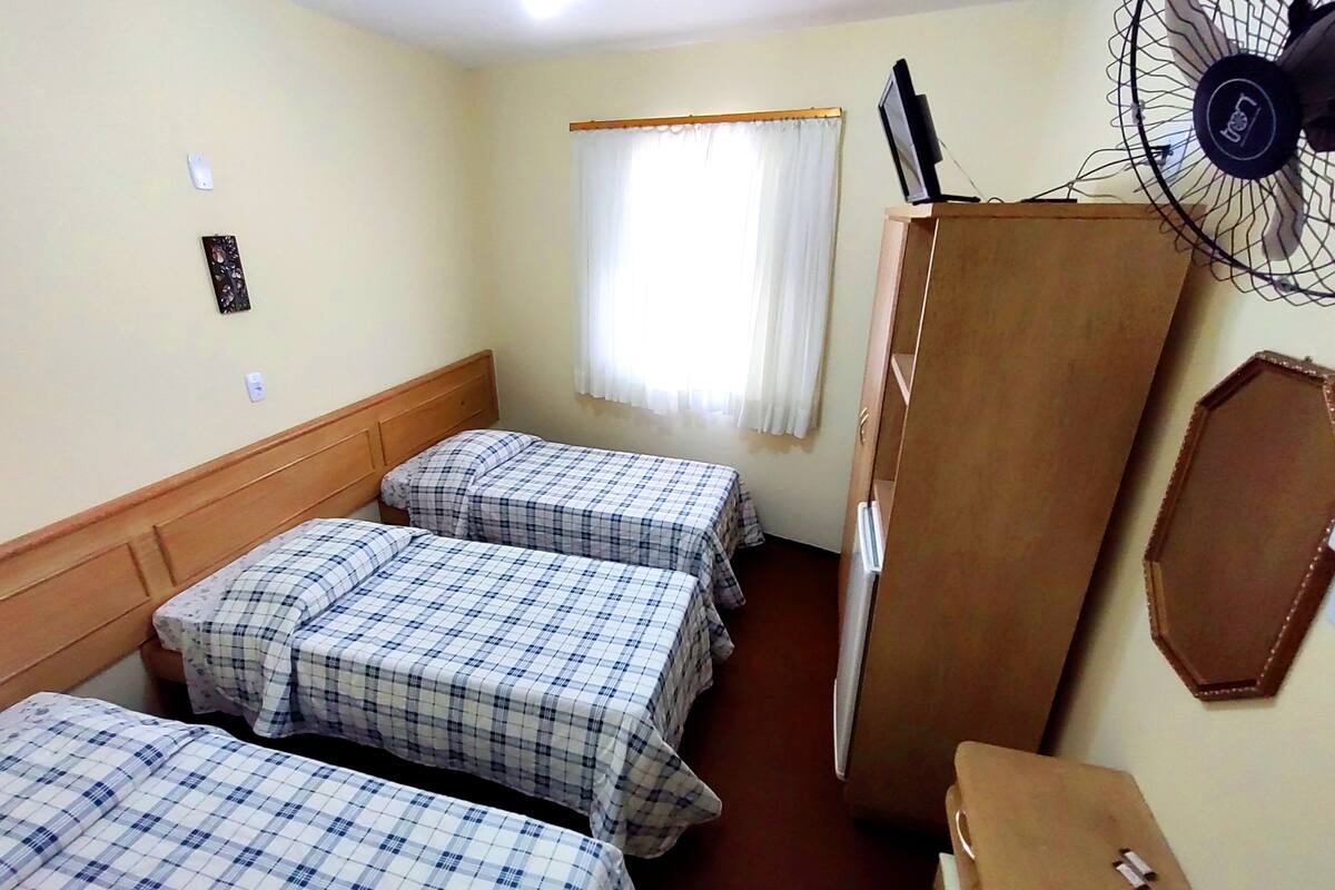 Una habitación con tres camas, un armario de madera, un ventilador y una ventana con cortinas.