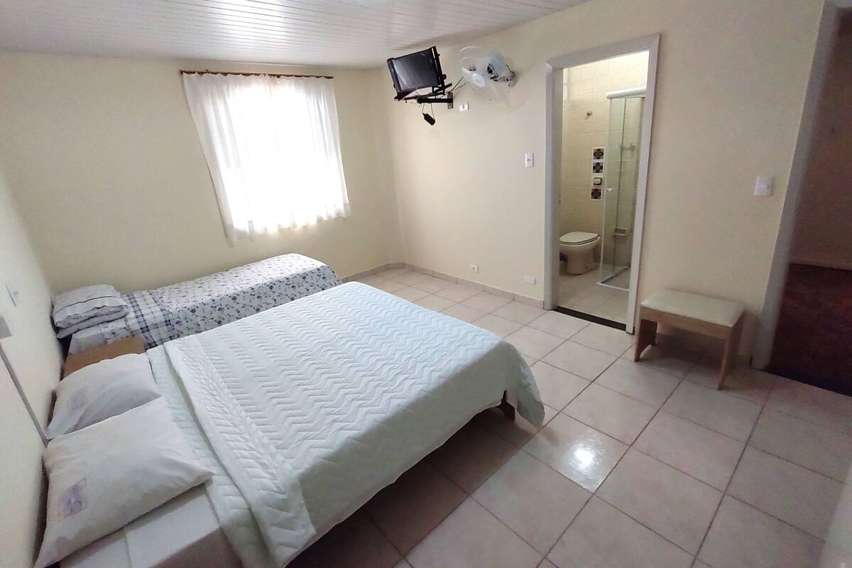 Un dormitorio con cama, una mesita de noche, un televisor montado en la pared y un baño visible a través de una puerta abierta.
