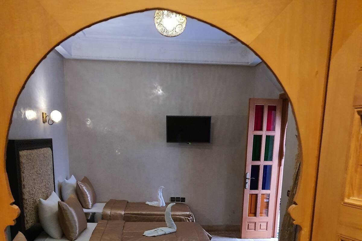 A hotel room with two beds, each with a brown bedspread and a towel shaped like a swan. A flat-screen TV is mounted on the wall, and a wooden door with colorful glass panels is visible.