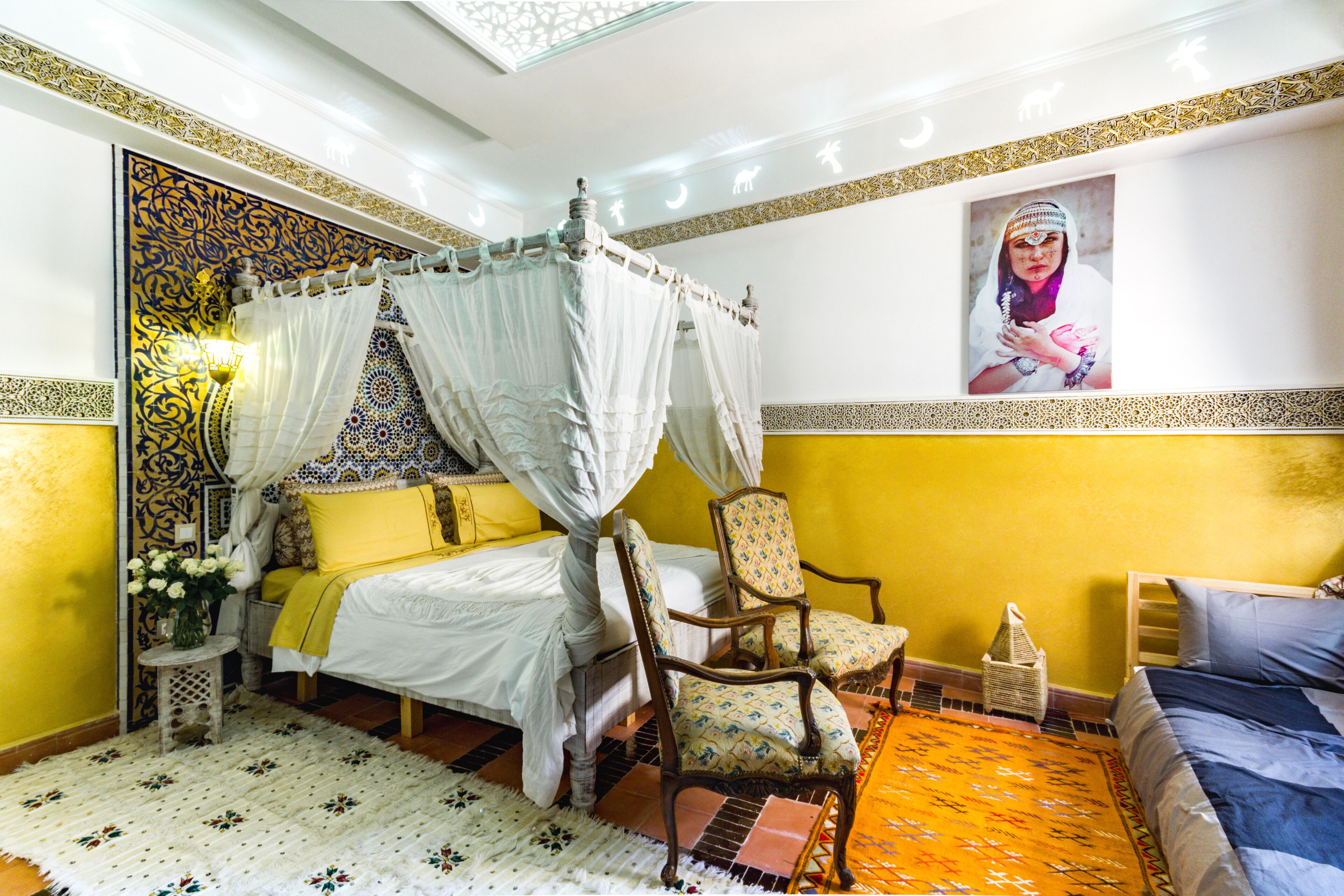 Une chambre avec un lit à baldaquin, une chaise, une petite table sur laquelle est posé un vase de fleurs, et un mur orné d’un motif décoratif.