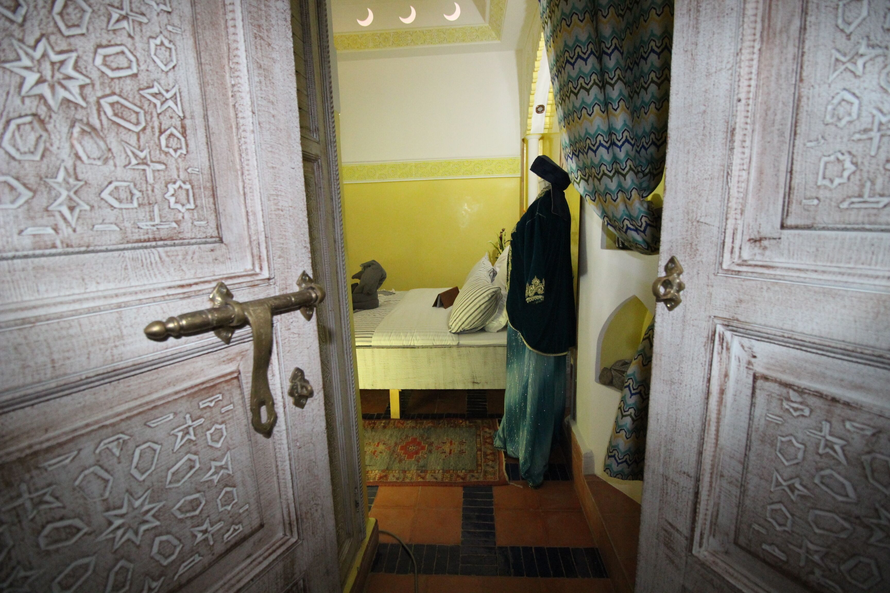 A room with a bed, a door with intricate patterns, a hanging garment, and a patterned rug.