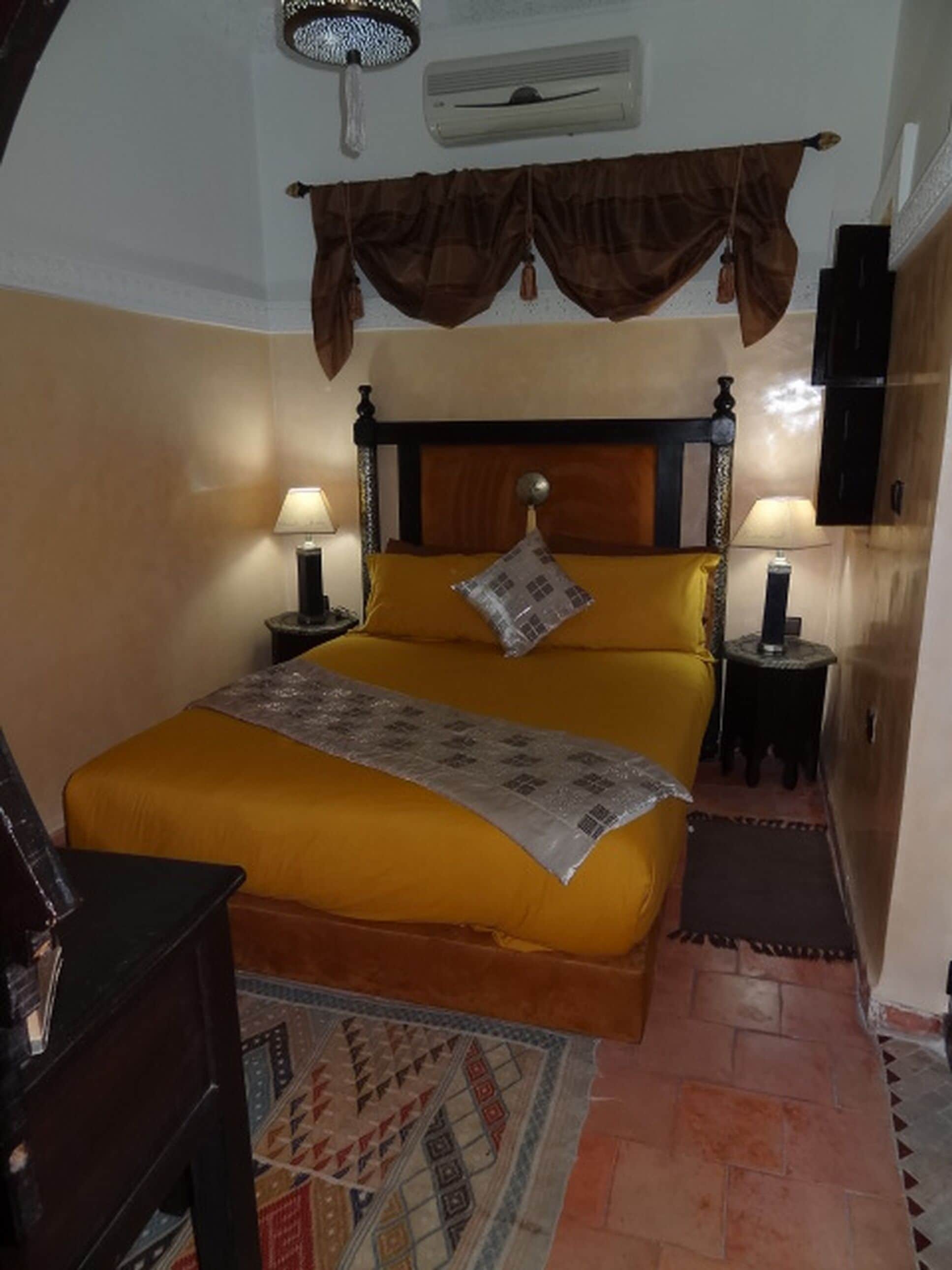 Un dormitorio con una cama grande, dos mesitas de noche con lámparas, un cabecero decorativo, un ventilador de techo y una alfombra estampada.
