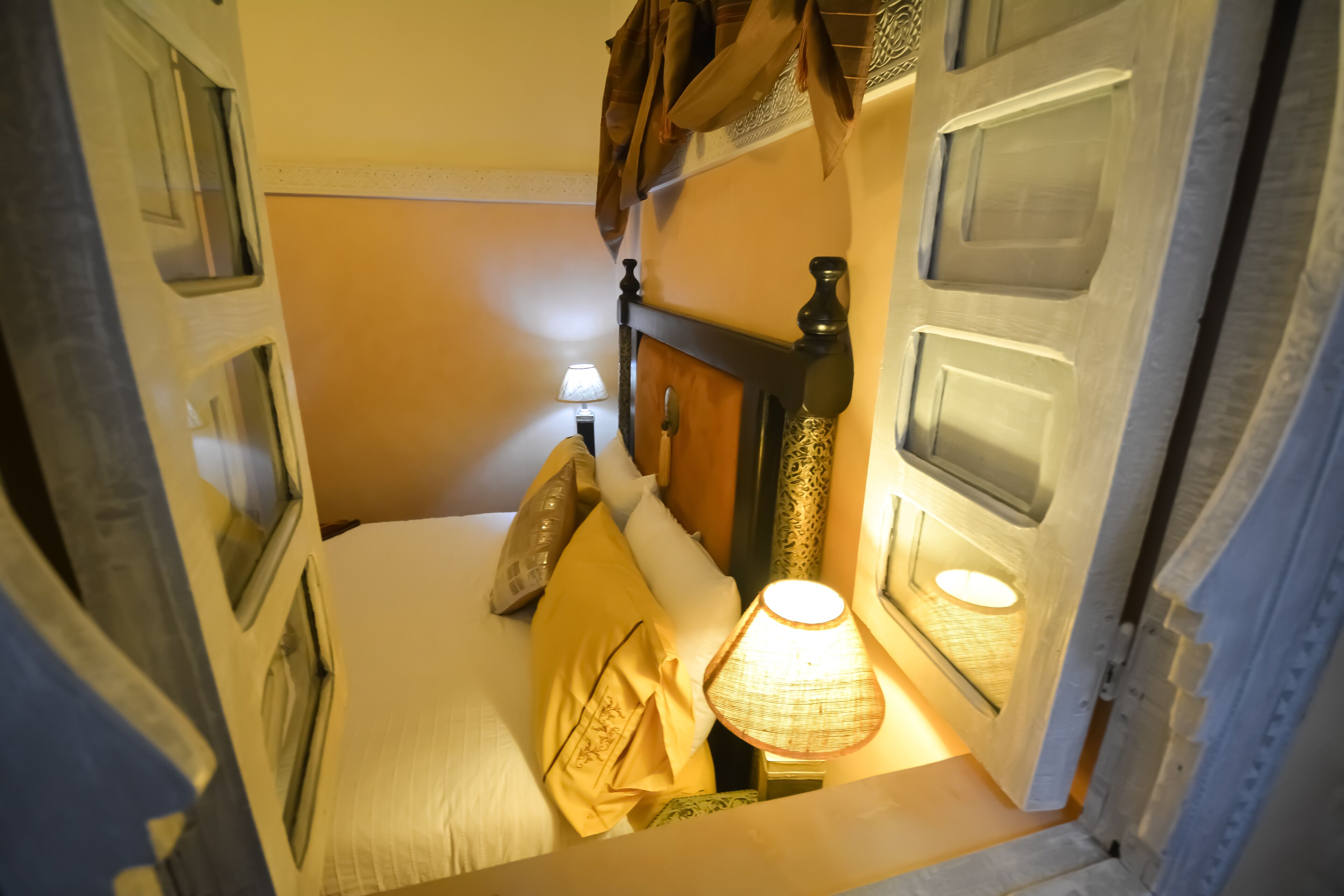 Una habitación de hotel estrecha con una cama, una mesita de noche con una lámpara y una ventana con cortinas.