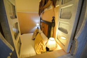 Una habitación de hotel estrecha con una cama, una mesita de noche con una lámpara y una ventana con cortinas.