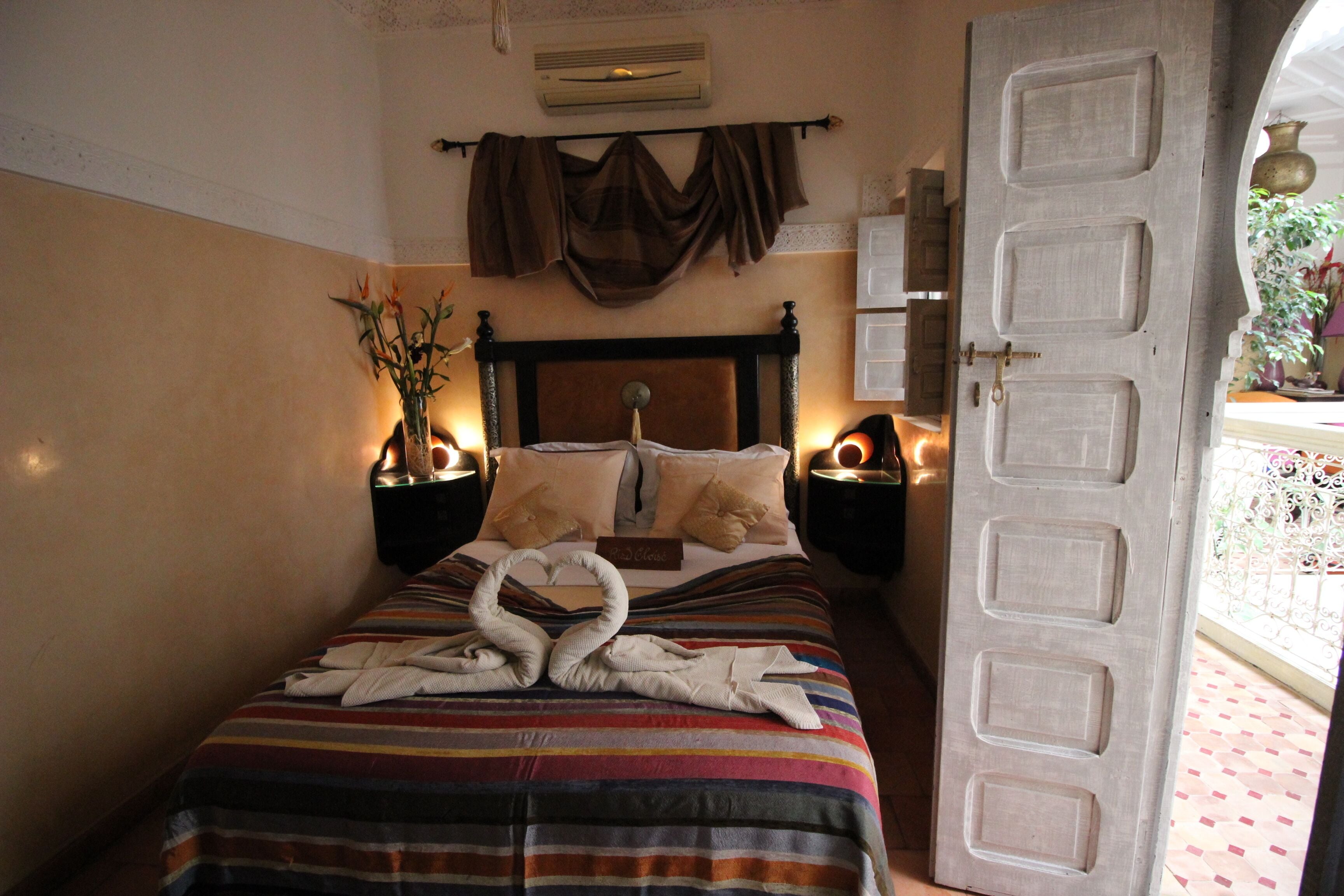 A hotel room with a bed featuring a striped bedspread, a headboard with a decorative drape, and a pair of towels arranged in a swan shape on the bed.
