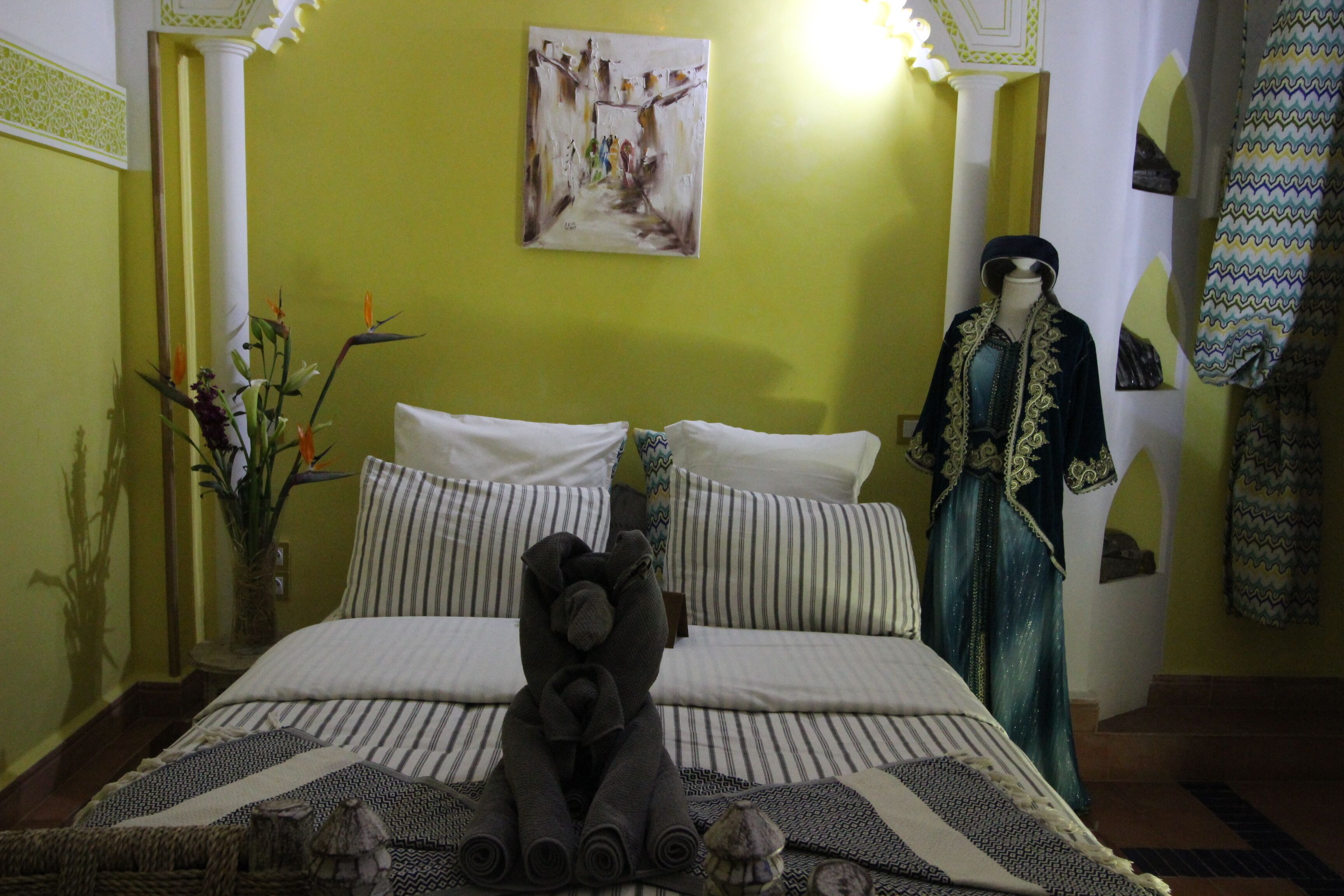 A hotel room with a bed, a decorative towel arrangement, a mannequin in traditional attire, and a painting on the wall.
