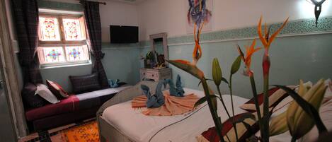 Una habitación con cama, sofá, ventana con cortinas, televisor montado en la pared y una planta decorativa.