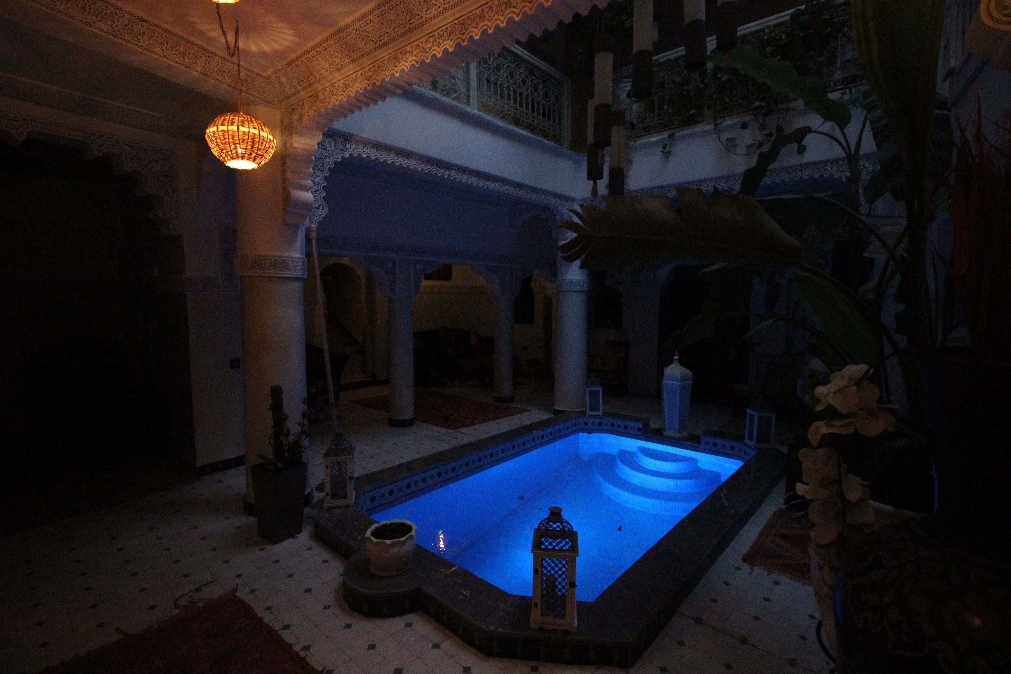 Ein spärlich beleuchteter Poolbereich im Innenbereich mit einer blau beleuchteten Wasserspielen, umgeben von aufwendigen architektonischen Details und Zimmerpflanzen.