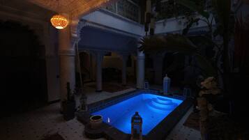 A dimly lit indoor pool area with a blue-lit water feature, surrounded by ornate architectural details and potted plants.