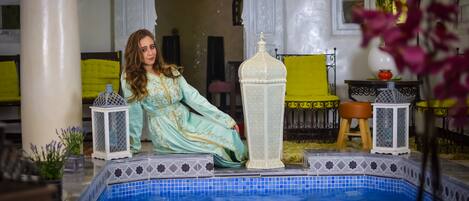 Zona de piscina con una persona vestida con traje tradicional, columnas blancas ornamentadas y una silla amarilla.