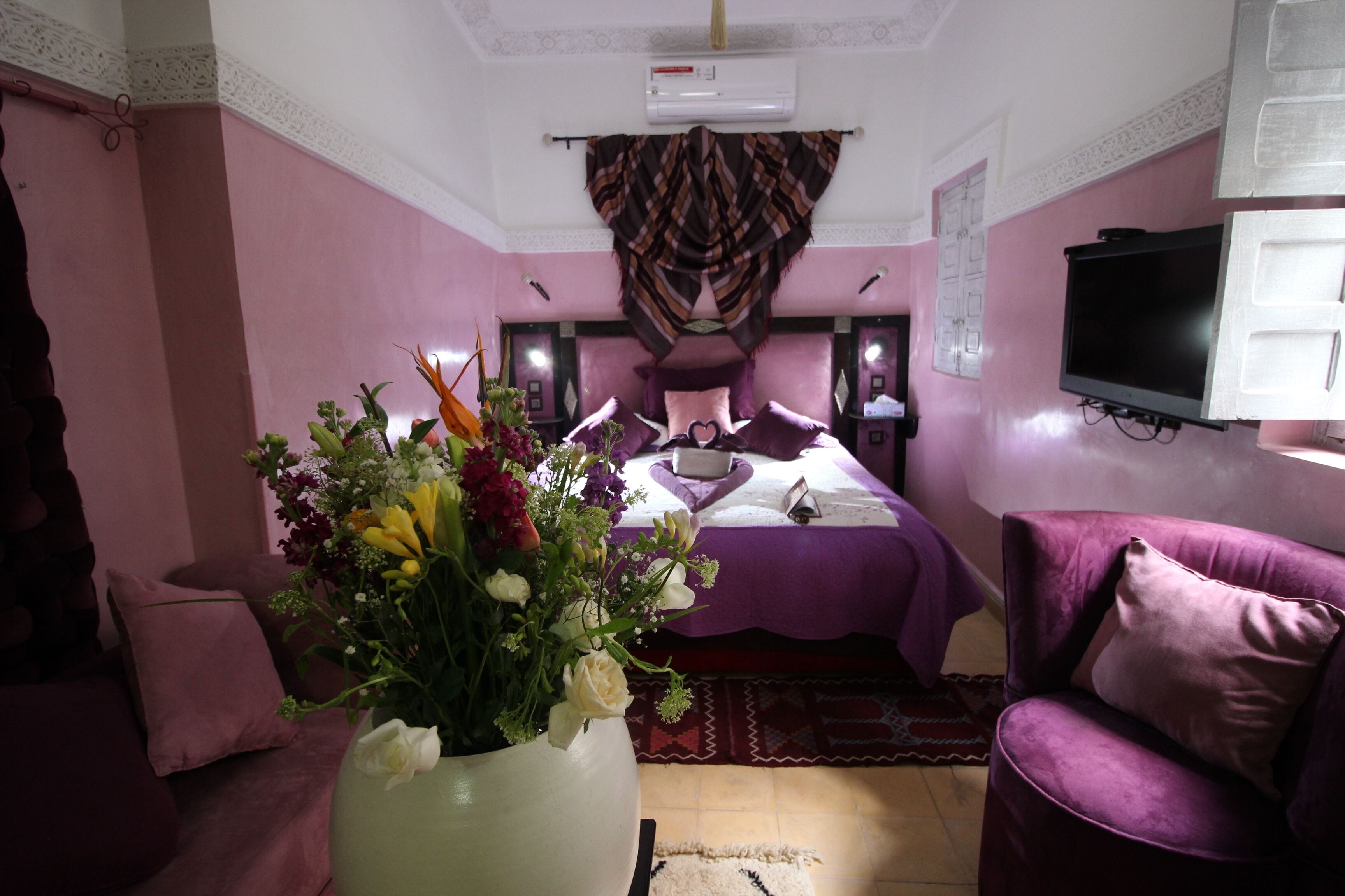 A bedroom with a bed, a TV, a purple sofa, and a floral arrangement.
