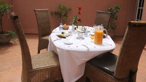A round dining table with a white tablecloth, surrounded by wicker chairs. There are potted plants, a red flower, and a pitcher of orange juice on the table. - Rooms Palace (Marrakech)