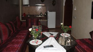 A hotel room with a living area featuring a sofa and a dining table set with a vase of flowers, a remote control, and a tray with glasses and a pitcher. - Rooms Palace (Marrakech)