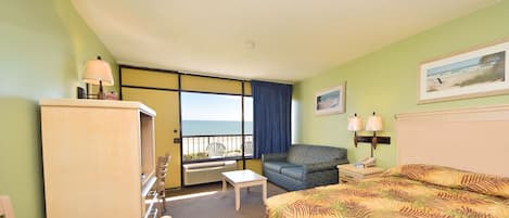 A hotel room with a bed, a desk, a chair, a small table, a sofa, a nightstand, a lamp, a picture on the wall, and a view of the ocean through a large window.