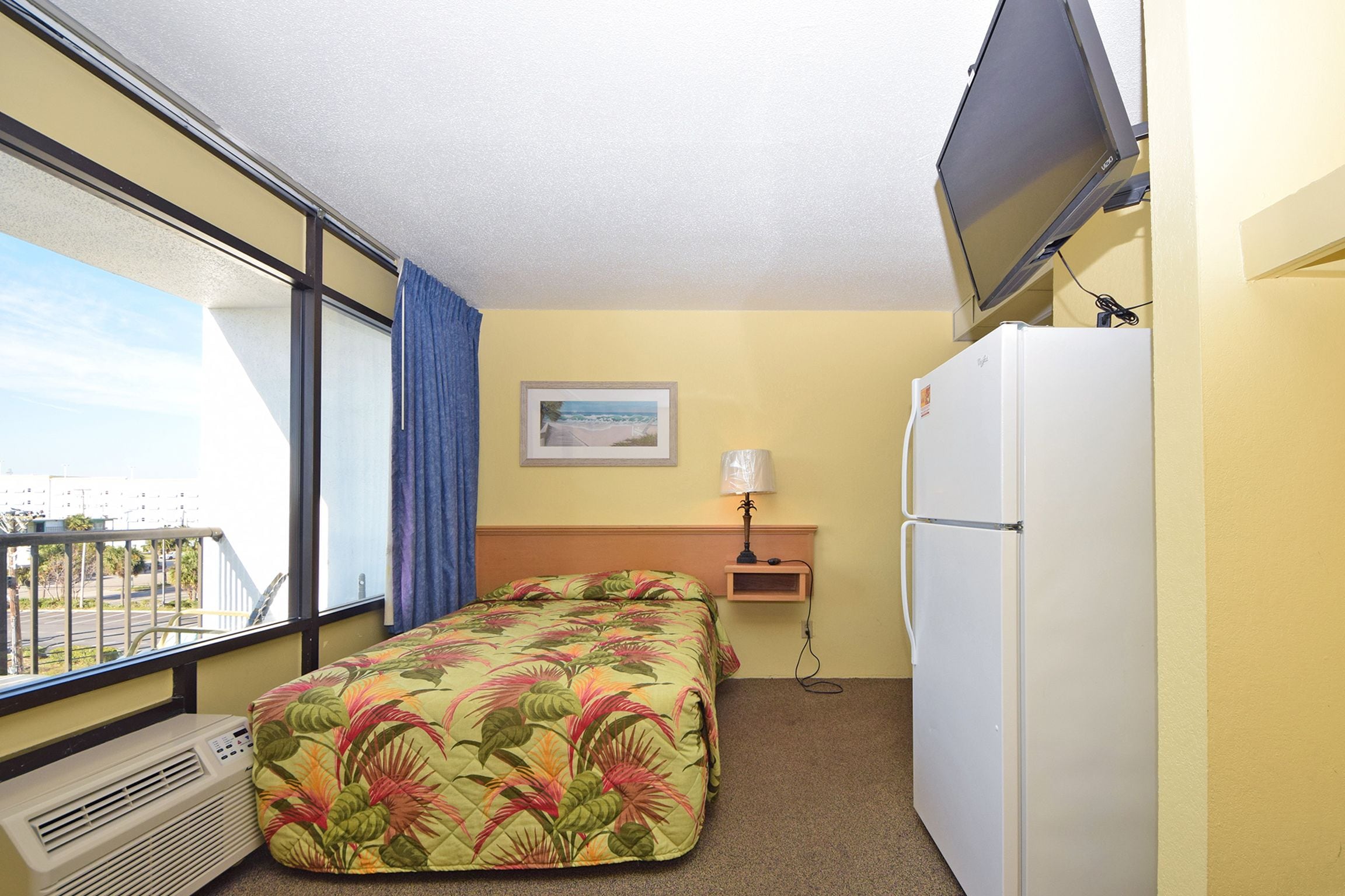 A hotel room with a bed, a refrigerator, a TV mounted on the wall, a window with a view, and a small table with a lamp.