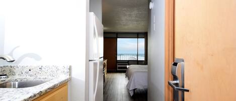 A narrow hallway with a kitchen area on the left, a bed with a white bedspread in the center, and a wooden door on the right.
