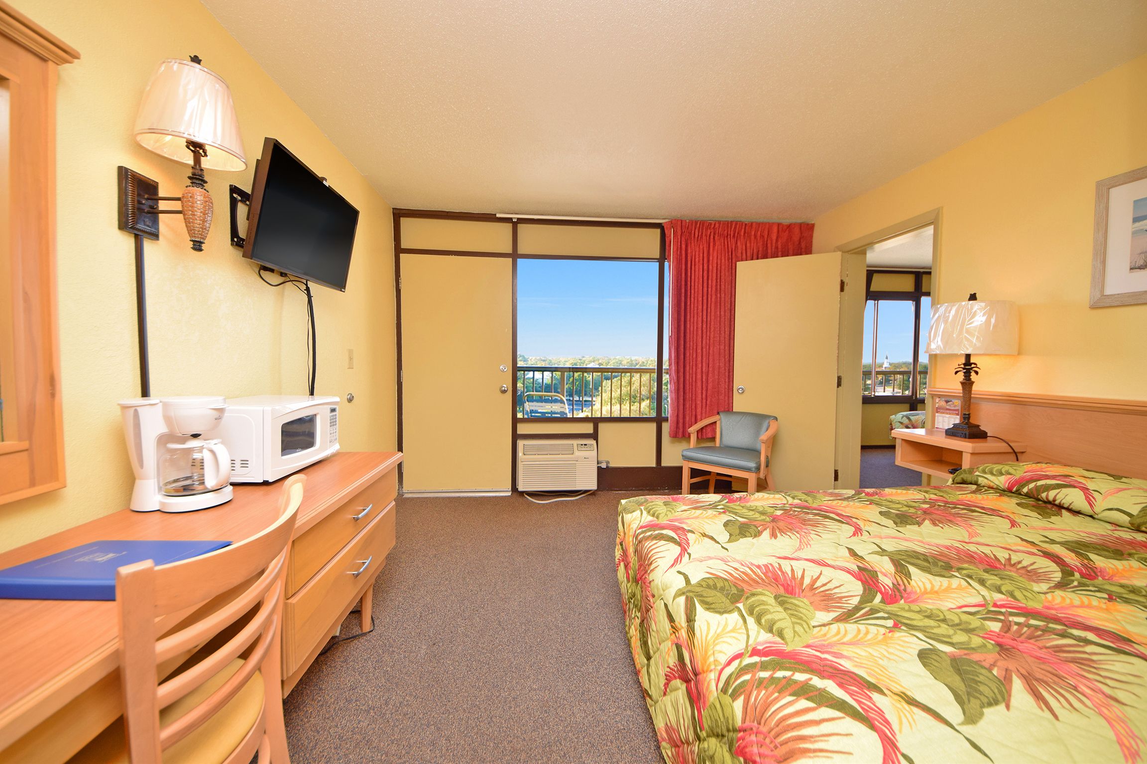 A hotel room with a bed, a desk with a coffee maker, a TV, and a view of the outside.