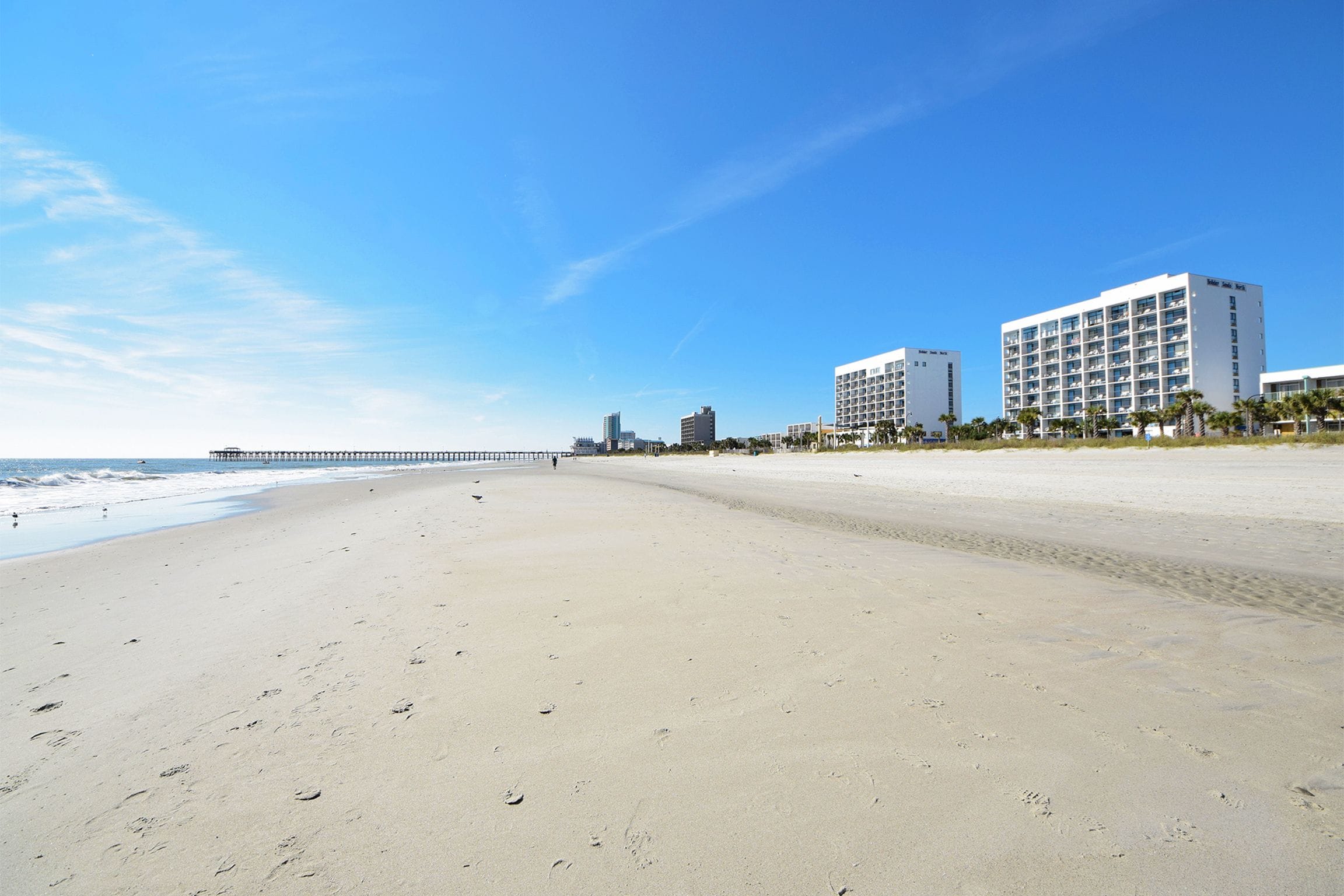 Une plage avec une jetée qui s’avance dans l’océan, un ciel bleu dégagé et un hôtel à plusieurs étages doté de balcons.
