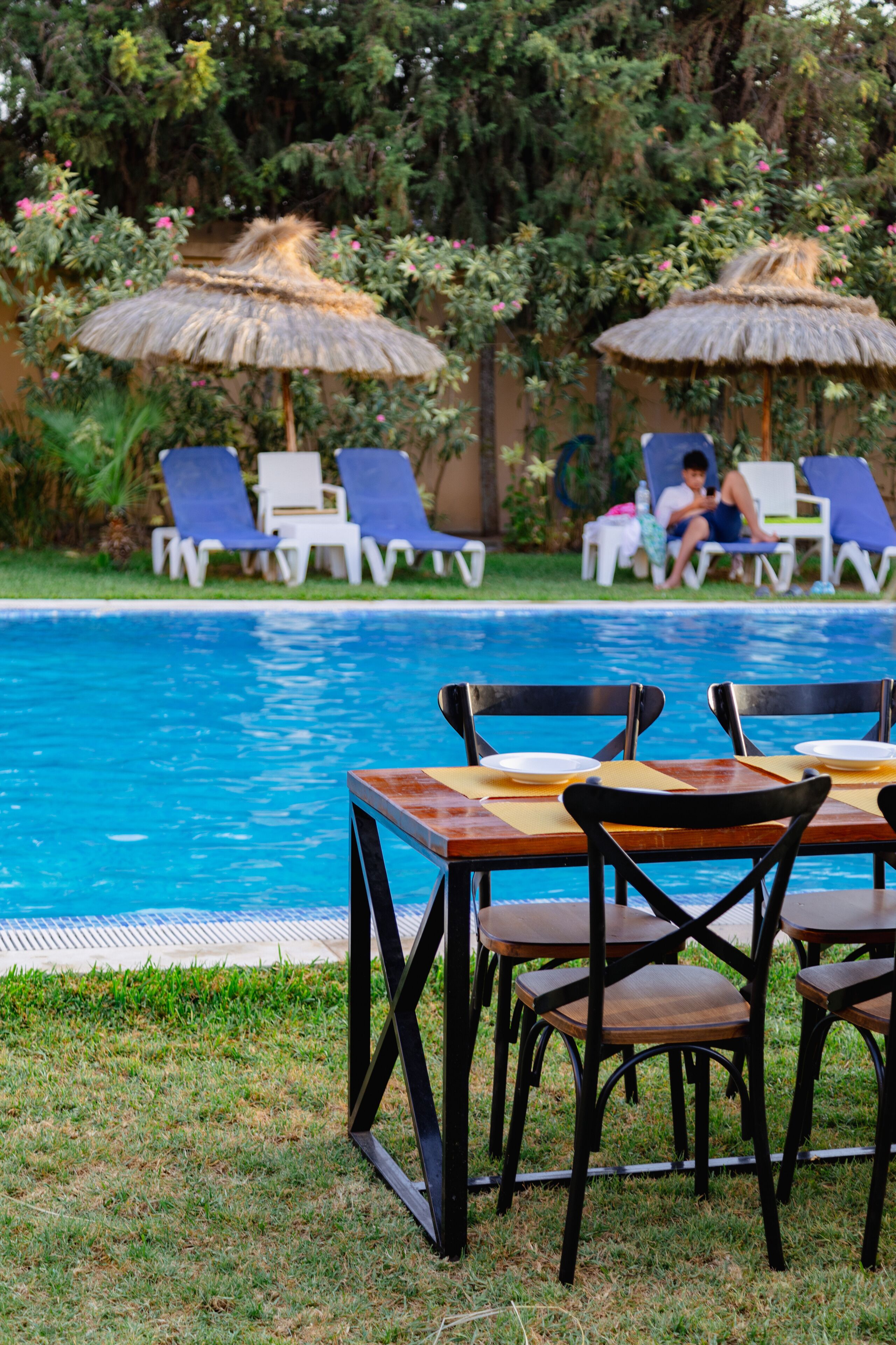 A poolside area with lounge chairs and tables, surrounded by greenery and a clear pool.