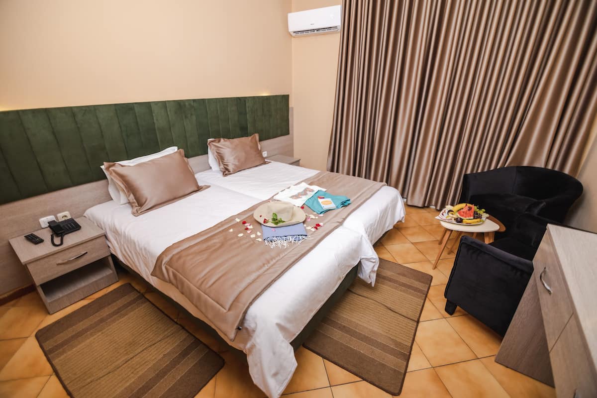A hotel room with a double bed, a nightstand with a phone and remote, a small table with fruits, and a black chair.