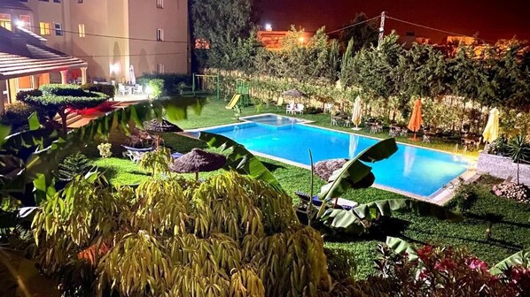 A well-lit outdoor swimming pool surrounded by neatly trimmed grass and tropical plants.