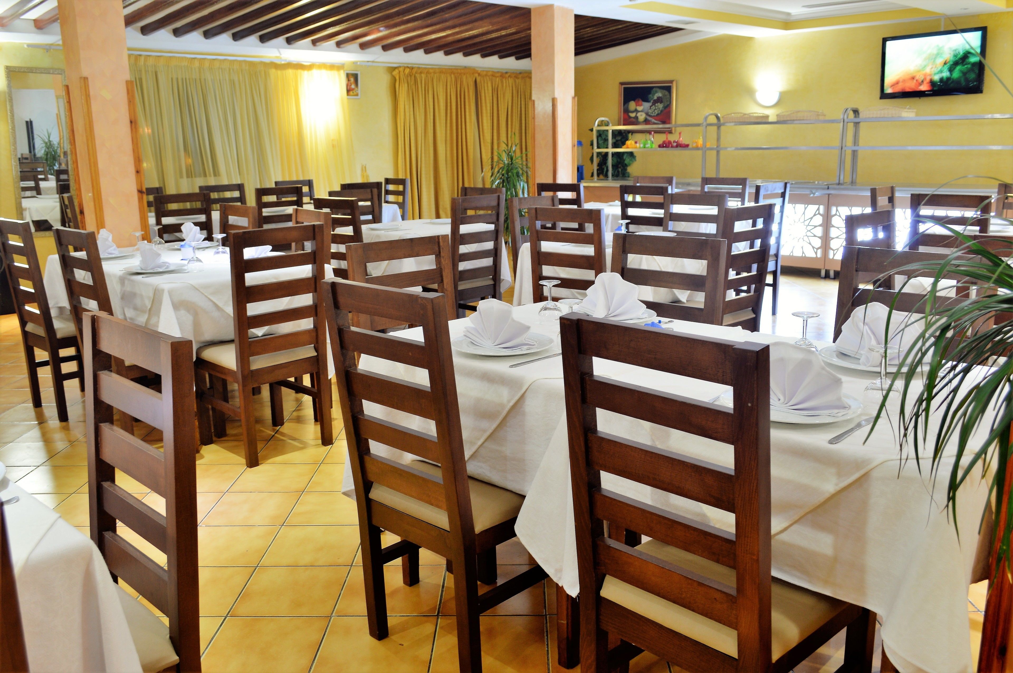 A restaurant with multiple tables and chairs, yellow curtains, and a television on the wall.