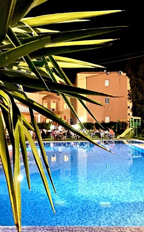 A swimming pool with a clear blue water surface, surrounded by palm leaves. In the background, there is a building with multiple windows and balconies.