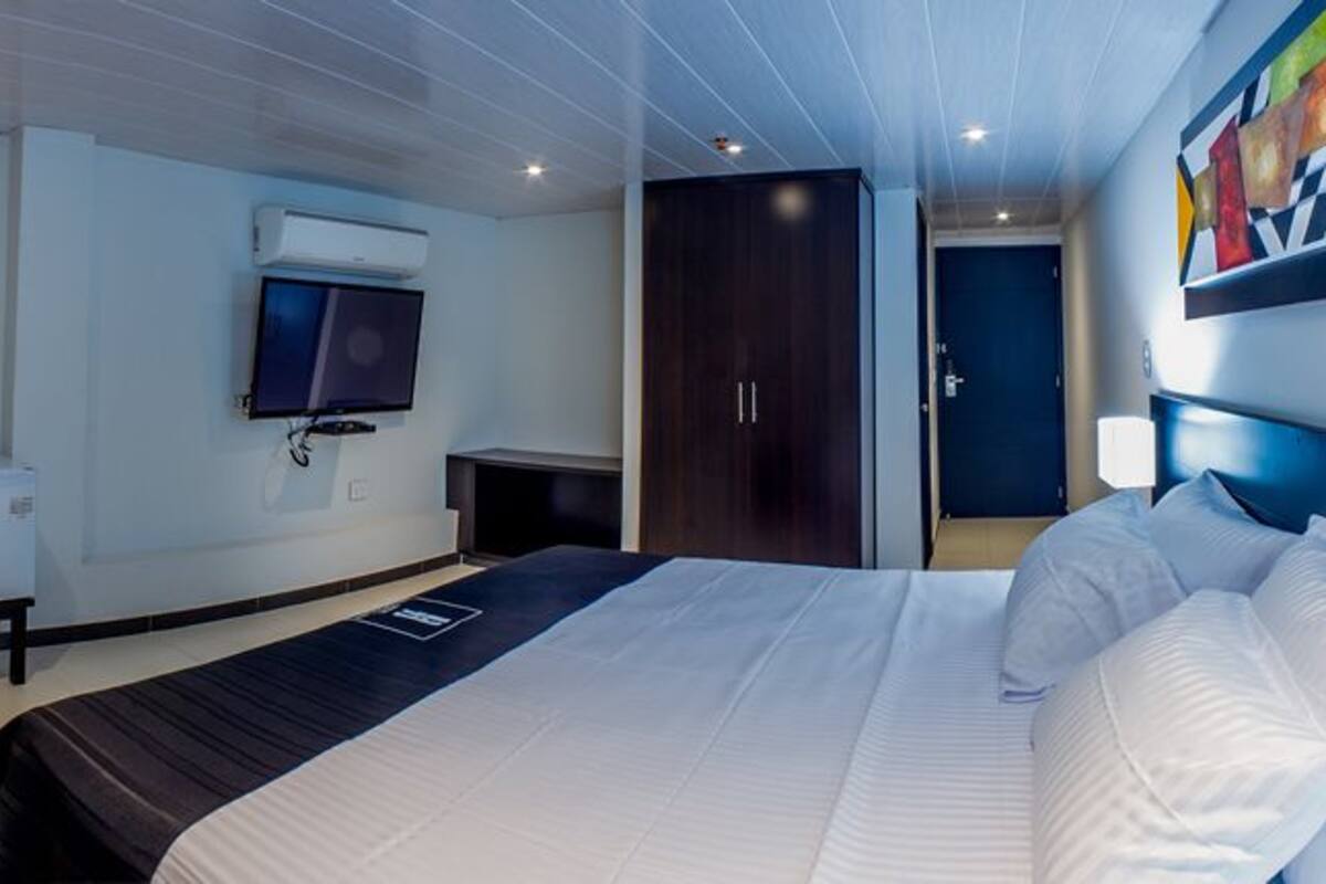 A modern hotel room with a bed, a desk, a TV mounted on the wall, and a door leading to another room.