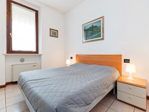Een slaapkamer met een bed, een nachtkastje met een lamp, een radiator, een raam met gordijnen, en twee ingelijste foto’s aan de muur.