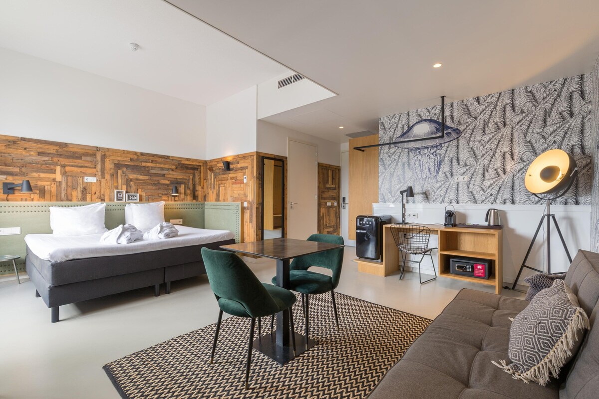 A modern hotel room with a bed, a dining table with chairs, a sofa, a desk with a chair, and a decorative wall.