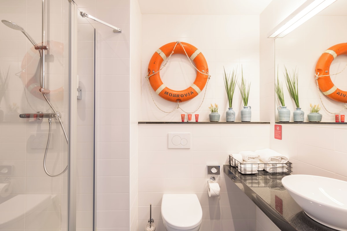 Un baño moderno con cabina de ducha de vidrio, un inodoro blanco, un lavamanos grande y dos anillos salvavidas naranjas en la pared.