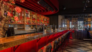 Una zona de bar con una barra larga, varias estanterías llenas de botellas y una fila de taburetes rojos.