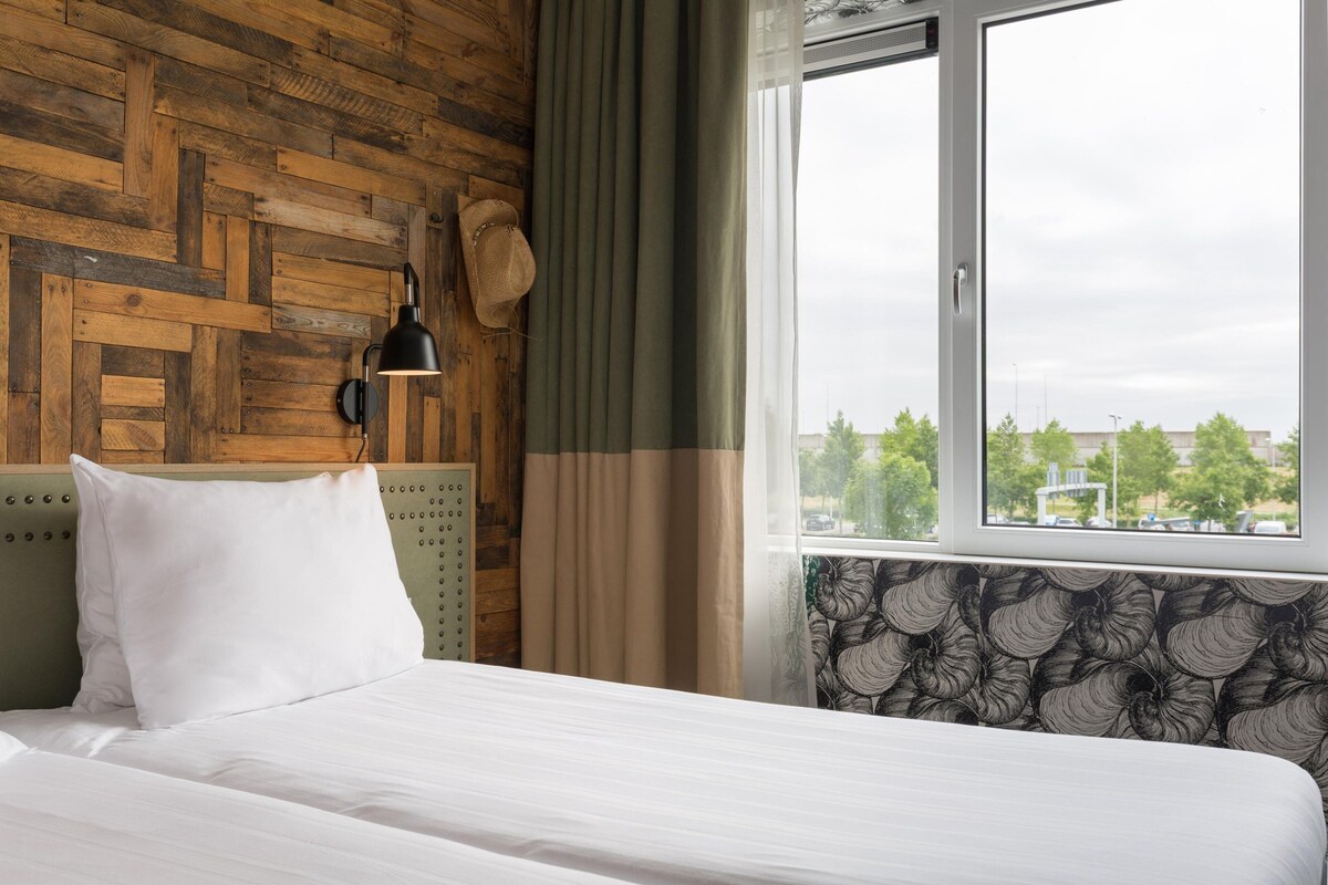 A bedroom with a wooden headboard, a bed with white linens, a black wall lamp, and a window with a view of trees and buildings.