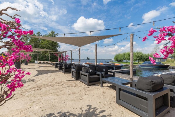 Un área de descanso junto a la playa con sillones reclinables y una pérgola, con vista a una masa de agua y barcos.
