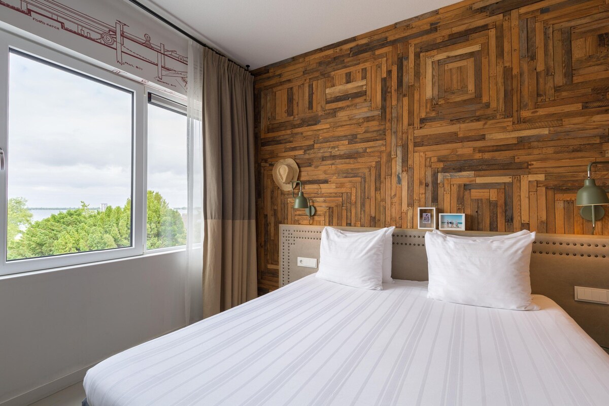 A hotel room with a large bed, wooden wall paneling, a view of trees through the window, and a curtain.