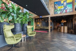 Um lobby com design moderno, apresentando uma planta grande, duas cadeiras verdes, piso de madeira e um mural colorido na parede.