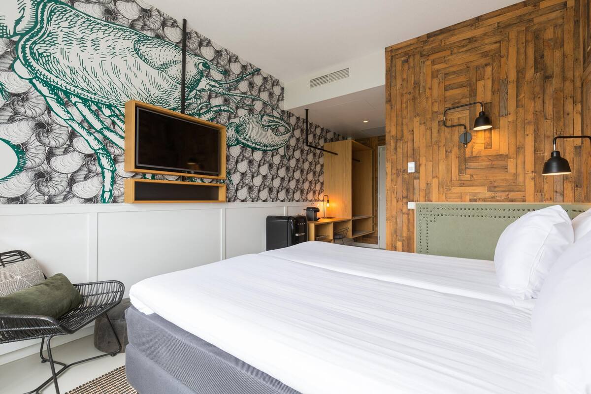 A modern hotel room with a large bed, a flat-screen TV mounted on the wall, a wooden accent wall, and a small kitchenette area.