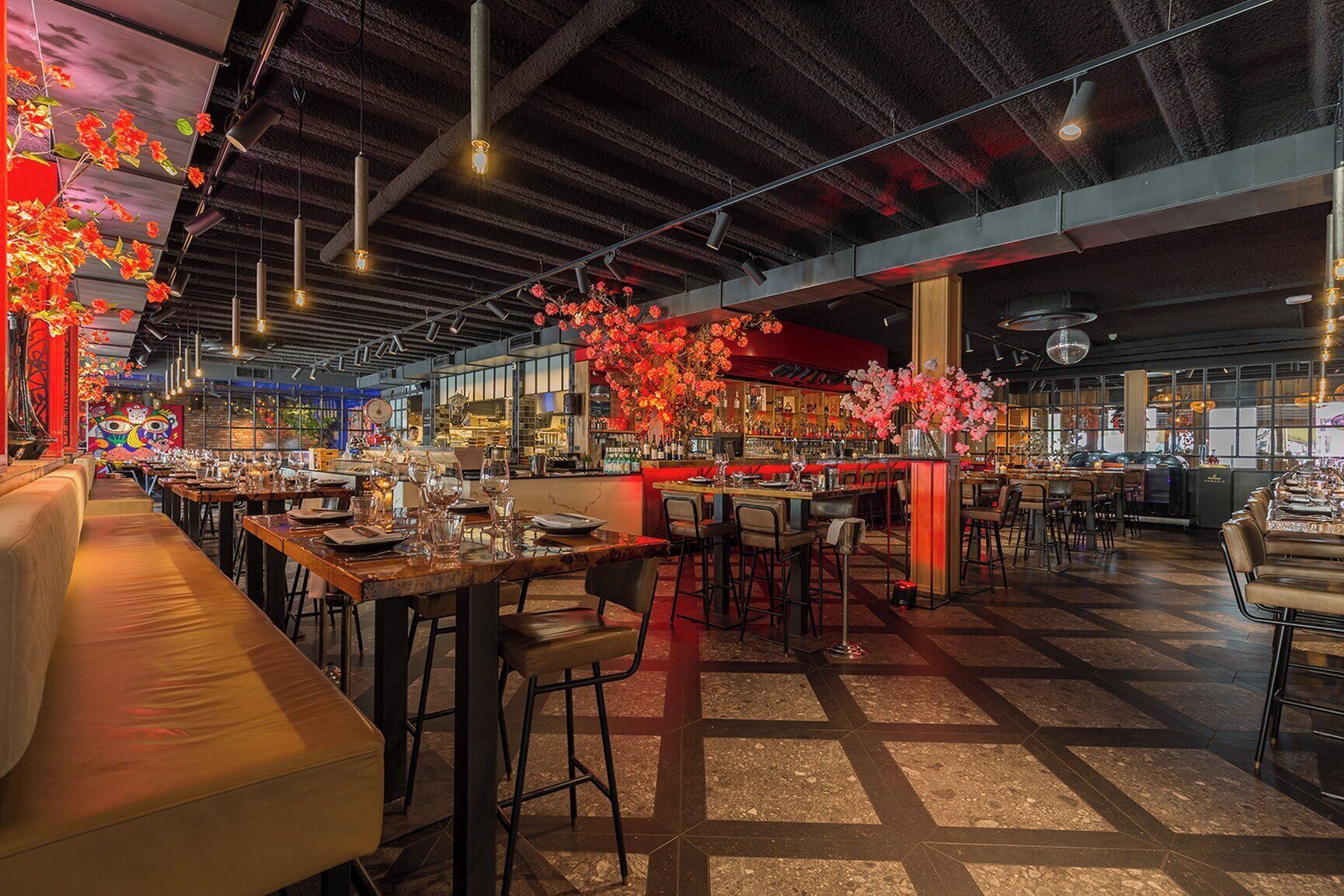 Un restaurant spacieux avec de hauts plafonds, des poutres apparentes et un mélange d’espaces bar et salle à manger. Les tables sont dressées avec des verres et des couverts, et des compositions florales ornent le comptoir du bar.