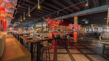 Un restaurant spacieux avec de hauts plafonds, des poutres apparentes et un mélange d’espaces bar et salle à manger. Les tables sont dressées avec des verres et des couverts, et des compositions florales ornent le comptoir du bar.