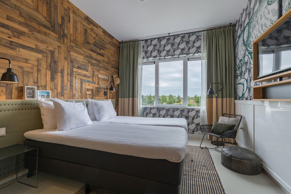 A bedroom with a wooden wall, a large bed, a window with curtains, a chair, and a desk.