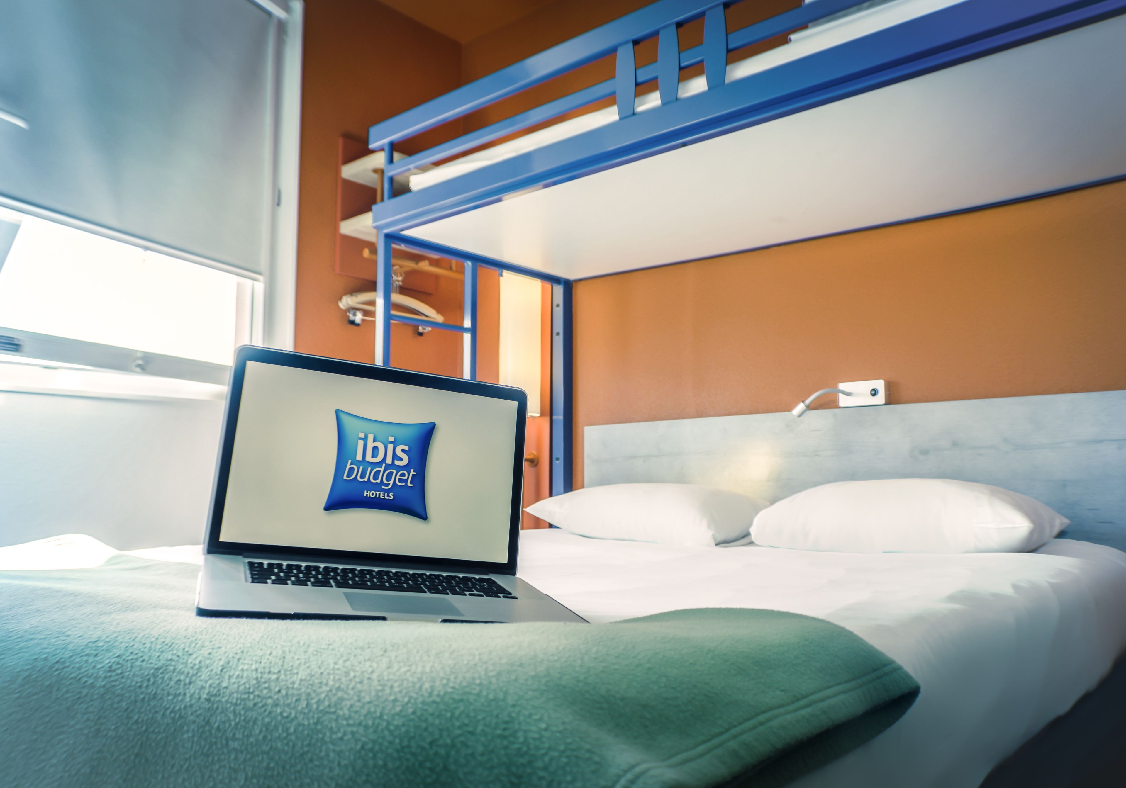 Una computadora portátil sobre una cama mostrando el logo del hotel ibis budget, una cama litera y una mesita de noche con una lámpara.