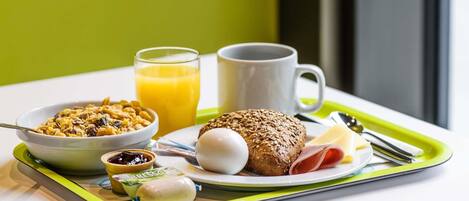Ein Frühstücksbrett mit Müsli, Saft, einer Tasse, einem Brötchen, einem gekochten Ei, einer Scheibe Schinken und einer kleinen Marmeladengläschen.