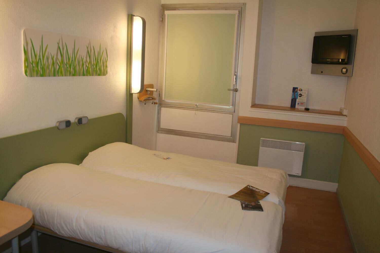 Ein Hotelzimmer mit zwei Einzelbetten, einem Nachttisch, einem Spiegel, einem Fernseher und einer Heizung.